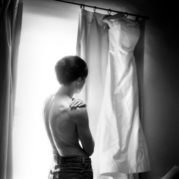black and white photo - view of of the back of the beautiful bride wearing only jeans and a french twist hairdo looking her hanging wedding dress - photo by New Mexico based wedding photographers Twin Lens